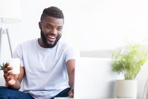 African American man sits at his desk, smiling brightly while looking at his laptop screen. He is holding a white mug of coffee and appears to be working from home, copy space photo