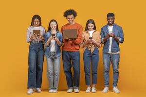 This image shows a group of five young adults standing in a row against a yellow background. They are all looking down at their devices, which include a tablet, a smartphone, and a laptop photo