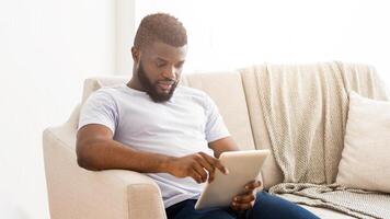 African American man is sitting on a couch in a living room. He is relaxed and using a tablet device. The room is brightly lit, and the man appears to be focused on the screen. photo