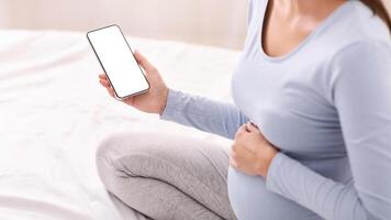 Pregnancy Apps. Unrecognizable Pregnant Girl Holding Cellphone With Blank Screen Sitting On Bed At Home. Cropped, Mockup photo