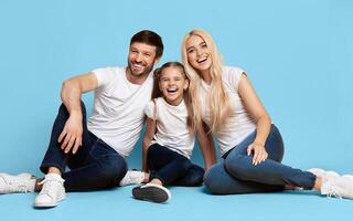A family of three, a father, a mother, and a daughter, are sitting on a blue background. They are all smiling and appear to be happy. The daughter is sitting in the middle with her parents photo