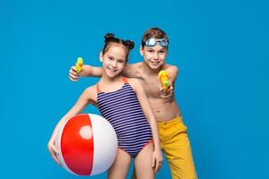 Two happy children are having fun with summer activities. A girl holds a beach ball while a boy aims a water gun. Both are dressed in swimwear, with a vibrant blue backdrop emphasizing lively mood. photo