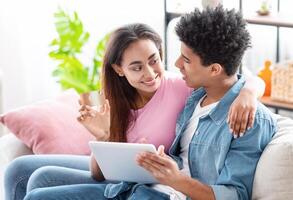 Black teen couple relaxing on a couch, both wearing casual attire, with the girl sitting on the guys lap. They are discussing something on a tablet computer, with the girl pointing at the screen. photo