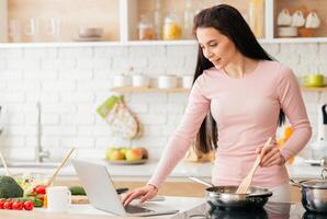 A woman is multitasking in the kitchen, cooking food while using a laptop for work or browsing the internet. She is standing near the stove, focusing on both tasks at hand. photo