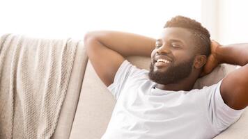 Black man relaxing on a couch. He is wearing a white t-shirt and has his arms behind his head. His eyes are closed and he is smiling. He appears to be enjoying a moment of peace and relaxation. photo