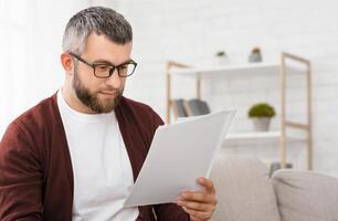 A man with glasses and a beard is intently reading papers while sitting on a sofa in a well-lit, modern home office. Shelves in the background hold various decor items photo