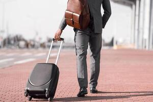Cropped of black businessman walking with rolling suitcase outside a modern building, ready for a business trip or travel. photo