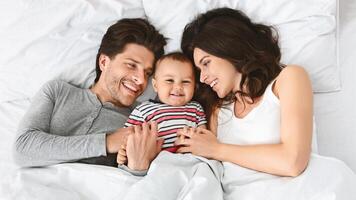 Cheerful family, a man and a woman, are lying in bed together, smiling at their baby. The baby is lying in the middle of the bed, between its parents, and is smiling back at them. photo