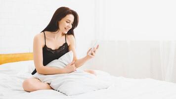 A woman is seated on a bed, holding a smartphone in her hands and looking at the screen. She appears focused on the device while sitting in a bedroom, copy space photo