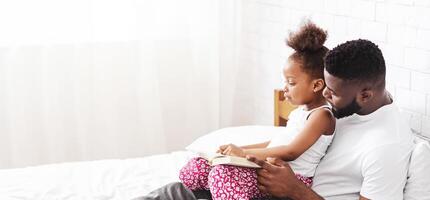 African American father sits on a white bed, reading to his daughter, who is seated on his lap, panorama with copy space photo