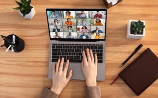 Lady uses a laptop for an online meeting with multiple participants. The laptop is open and the screen shows a grid of tiles of various people, all in a virtual meeting, top view photo