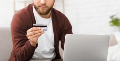 A man is pictured holding a credit card in one hand and looking at a laptop screen with a focused expression. He appears to be making an online purchase or managing his finances, cropped photo