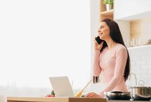 A woman is multitasking by talking on a phone and using a laptop simultaneously. She appears focused and engaged in both activities. photo