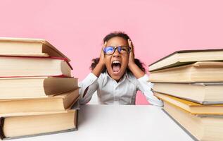 School Overload. African American Schoolgirl Shouting Clutching Head Sitting Between Two Book Stacks Over Yellow Studio Background. photo
