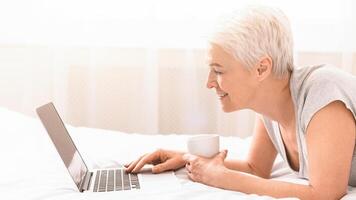 Senior woman is laying on a bed while using a laptop. She appears focused on the screen, with her hands typing or scrolling. The room is dimly lit, creating a cozy atmosphere, side view photo
