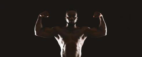 A muscular man flexes his biceps in a dark studio setting, with only his body illuminated. photo