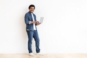 African American man is standing in front of a plain white wall, holding a laptop computer in his hands. He appears focused on the screen, perhaps working or browsing the internet. photo