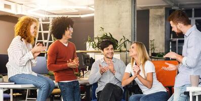 A team of five diverse coworkers are gathered in a bright, contemporary office setting, clapping and enjoying a moment of celebration. Their expressions reflect happiness and camaraderie photo