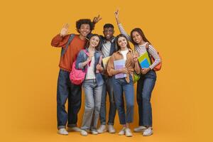 This image shows a group of diverse students smiling and posing together in front of a bright yellow background. They are all holding books and wearing backpacks photo