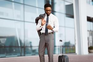 An executive black man with luggage checking phone outside a sleek airport. The setting is modern and busy, reflecting the life of a business traveler. photo