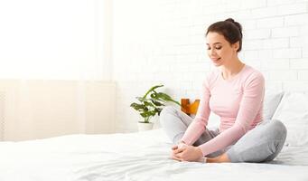 A woman is sitting on a bed with her legs crossed, displaying a relaxed posture. She appears to be comfortable and engrossed in a quiet moment, stretching, copy space photo