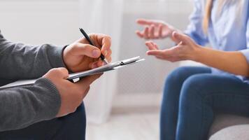 A close-up view of a professional taking notes on a clipboard while a client gestures with their hands, likely explaining a situation or expressing concerns, cropped photo