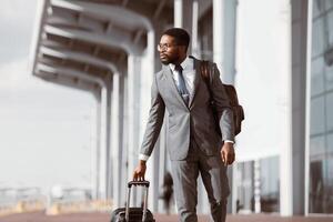 Black businessman walking with rolling suitcase outside a modern airport, ready for a business trip or travel. photo