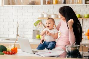 A mother and baby share a joyful moment in a modern kitchen. The baby sits on the counter while the mother looks at a laptop, both smiling and engaged. photo