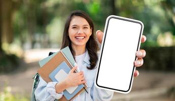 A young woman, likely a student, smiles brightly as she holds a smartphone with a blank screen. She is outdoors in a park-like setting, and her hand is outstretched, presenting the phone to the viewer photo