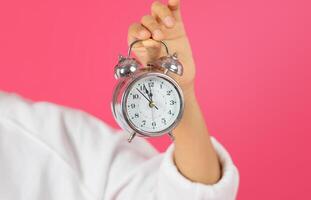 A hand is holding a silver alarm clock with the hands at 12 o'clock. The clock has a white face with black numbers and hands. The background is a bright pink, cropped photo