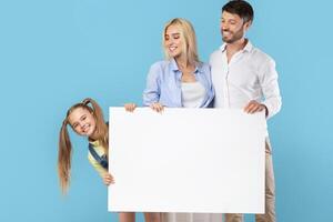 A young family, including a father, mother, and daughter, stand against a blue backdrop holding a large blank sign, looks happy and excited to show off the blank sign, perfect for adding your message photo