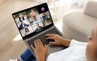 Black lady participates in a virtual meeting, engaging with a group of nine diverse professionals through a laptop in a bright, modern home setting. The screen displays smiling faces photo