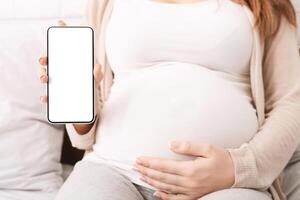 Modern technologies and pregnancy. Pregnant woman showing cellphone with empty display at home, mobile application for expectant ladies, mockup photo
