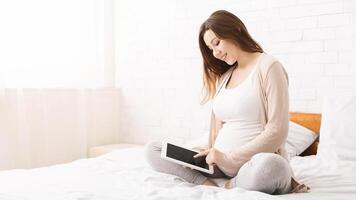 A pregnant woman is sitting cross-legged on a bed and smiling while using a tablet, creating a peaceful atmosphere as she enjoys her time connected to her device, copy space photo