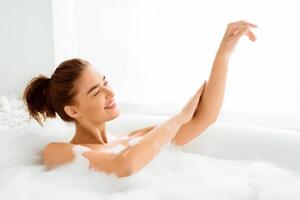 Beautiful woman enjoying pleasant bath with foam, applying cream on hands photo
