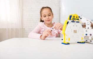 A young girl sits at a white table, focused on assembling a robot kit. She holds a small wire in her hand, carefully connecting it to the robots wiring. The robot is made of white plastic photo