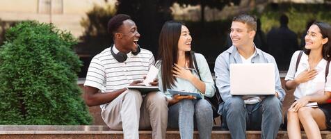 Break Between Classes. Happy group of students sitting on bench at university campus chatting and laughing photo