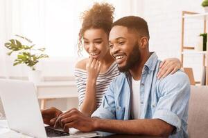 African American man and woman are seated on a couch, both focused on a laptop placed in front of them, engaged in whatever is on the screen, with expressions of concentration and interest. photo