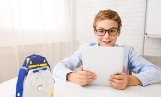 A young boy with glasses sits at a desk and uses a tablet. He looks intently at the screen, smiling. There is a small robot toy on the desk in front of him. photo