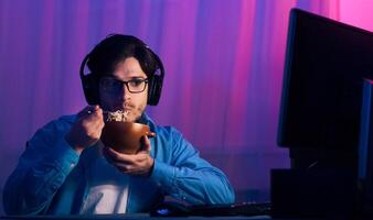 A young man sits in front of a computer screen, wearing headphones, while eating a bowl of noodles. He appears to be gaming at night, with colorful lights illuminating the background. photo