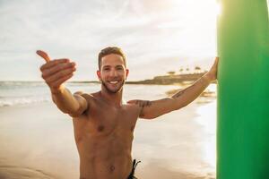 A young man stands on a sandy beach at sunset, holding a surfboard with his left hand and giving a thumbs up with his right. He is smiling and appears to be happy to be on the beach. photo