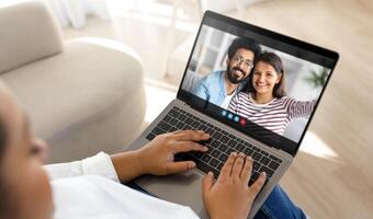 Black lady person sits on a couch at home, using a laptop computer to chat with a couple. The person is typing on the laptop, while the couple smiles and waves at the camera. photo
