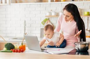 A mother is trying to work on her laptop in the kitchen while holding her curious baby. Various vegetables and notebook are on the counter, indicating attempt to balance work and parenting duties. photo
