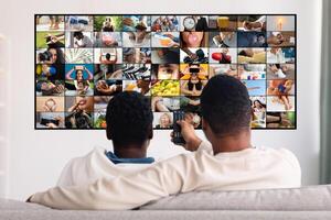 Two men relax on a couch in their home watching television. They are both wearing casual clothing and appear to be enjoying their time together. Television screen displays a montage of various images photo