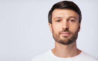 A man with a beard looks directly at the camera against a white background. He is wearing a white t-shirt. photo
