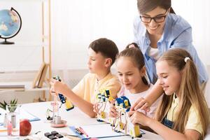 A group of elementary school students are working on a robotics project in a classroom setting. They are building robots with the help of their teacher, who is smiling and guiding them. photo