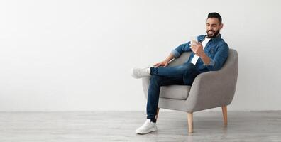 Cheerful young Arab man having online business meeting on smartphone, sitting in cozy armchair near white wall, banner design with free space. Eastern guy communicating remotely on mobile device photo