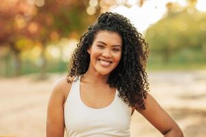 A young woman with curly hair smiles confidently while standing in a sunlit park. The background features trees and a warm, inviting atmosphere typical of the late afternoon. photo