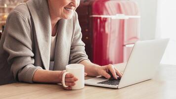 A smiling senior woman sits at a kitchen table, using a laptop computer. She is holding a white coffee mug in her left hand and her right hand is typing on the keyboard, cropped photo