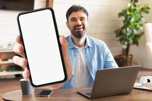 A man smiles while holding a smartphone with a blank screen, seated at a desk in a cozy home office with a laptop and warm decor around him. photo